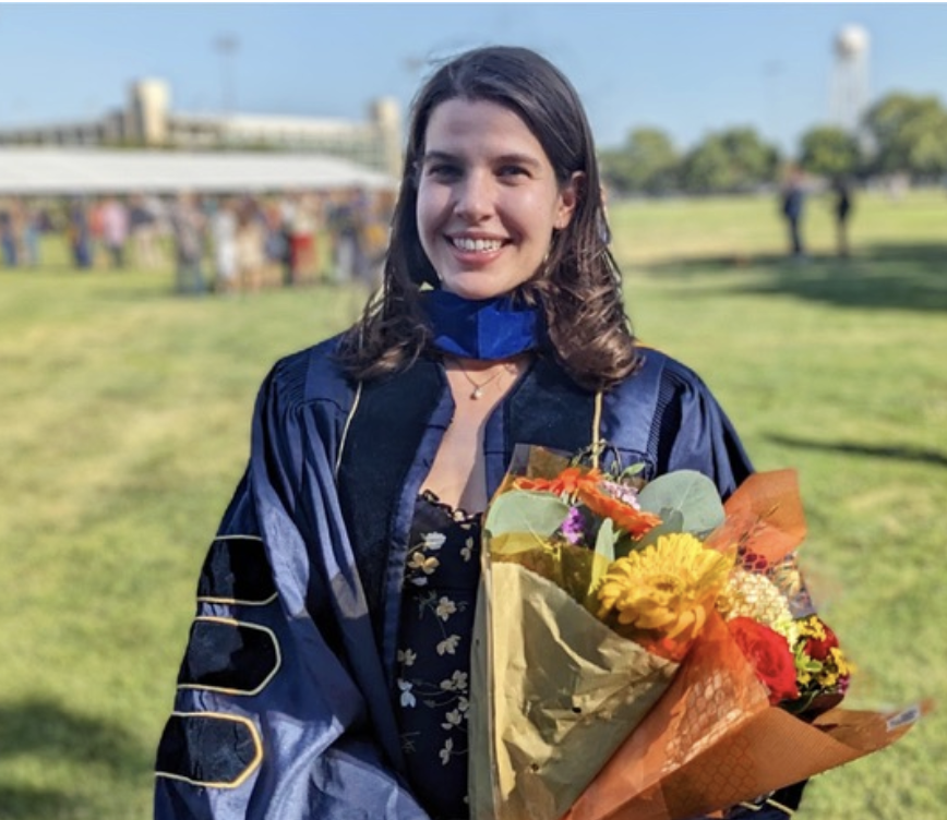 Ava Bindas '17 in a graduation gown, holding a bouquet of flowers outdoors on a sunny day.